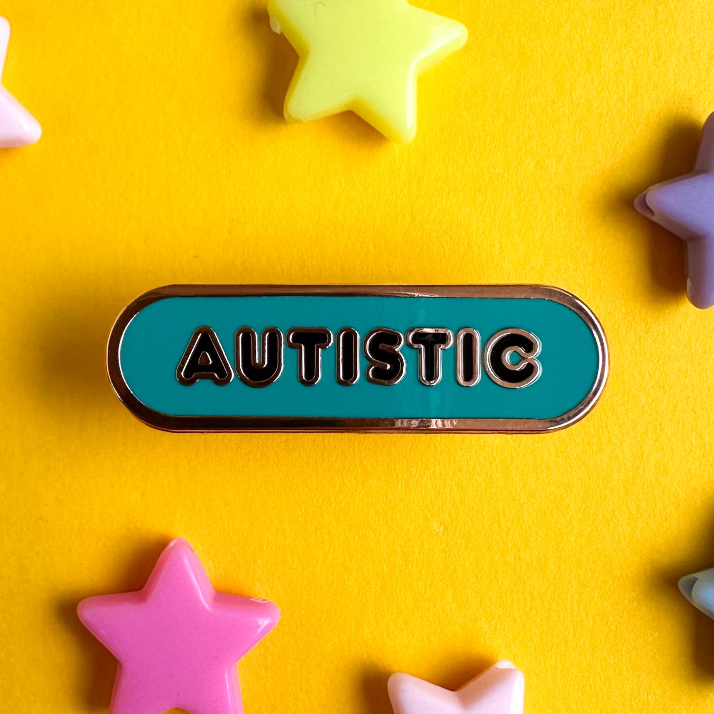 An enamel pin shaped like a bandaid that reads "Autistic" in black bubble letters, the background of the pin is teal. The pin is sitting on a piece of yellow paper that has pastel plastic star bead around it. 