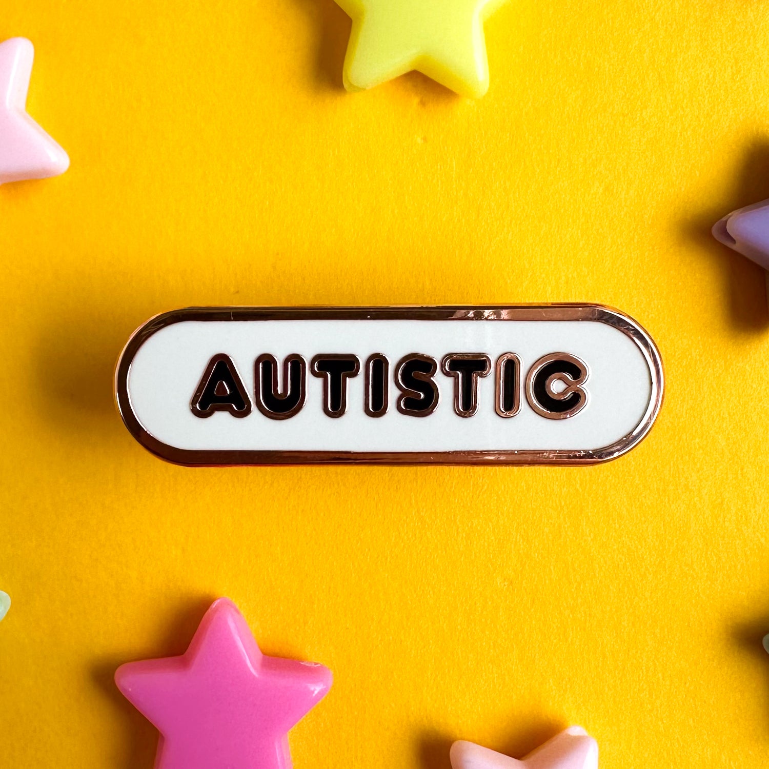 An oval shaped white pin with black bubble letters that read "Autistic" on it. The pin is on a yellow paper background with plastic star beads around it. 