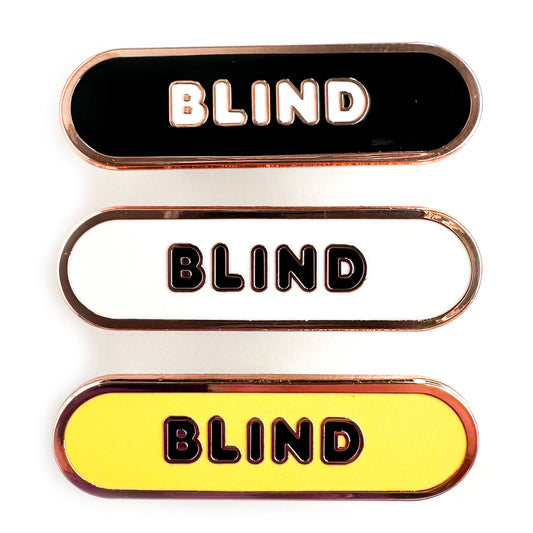 Three enamel pins in the shape of a capsule that all have bubble letters that read "Blind" the top pin has a black background, the middle is white, the bottom is yellow. 