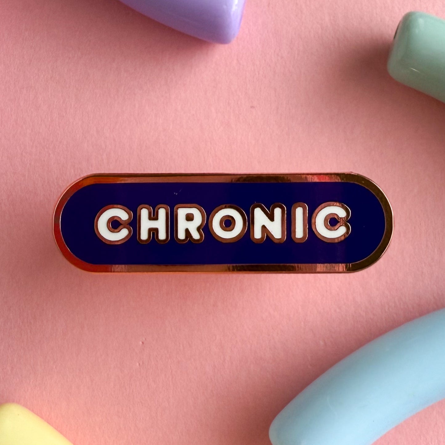 A bandaid shaped pin with the text CHRONIC in bubble letters. The background is navy blue and the text is white. The pin is on a pink background.