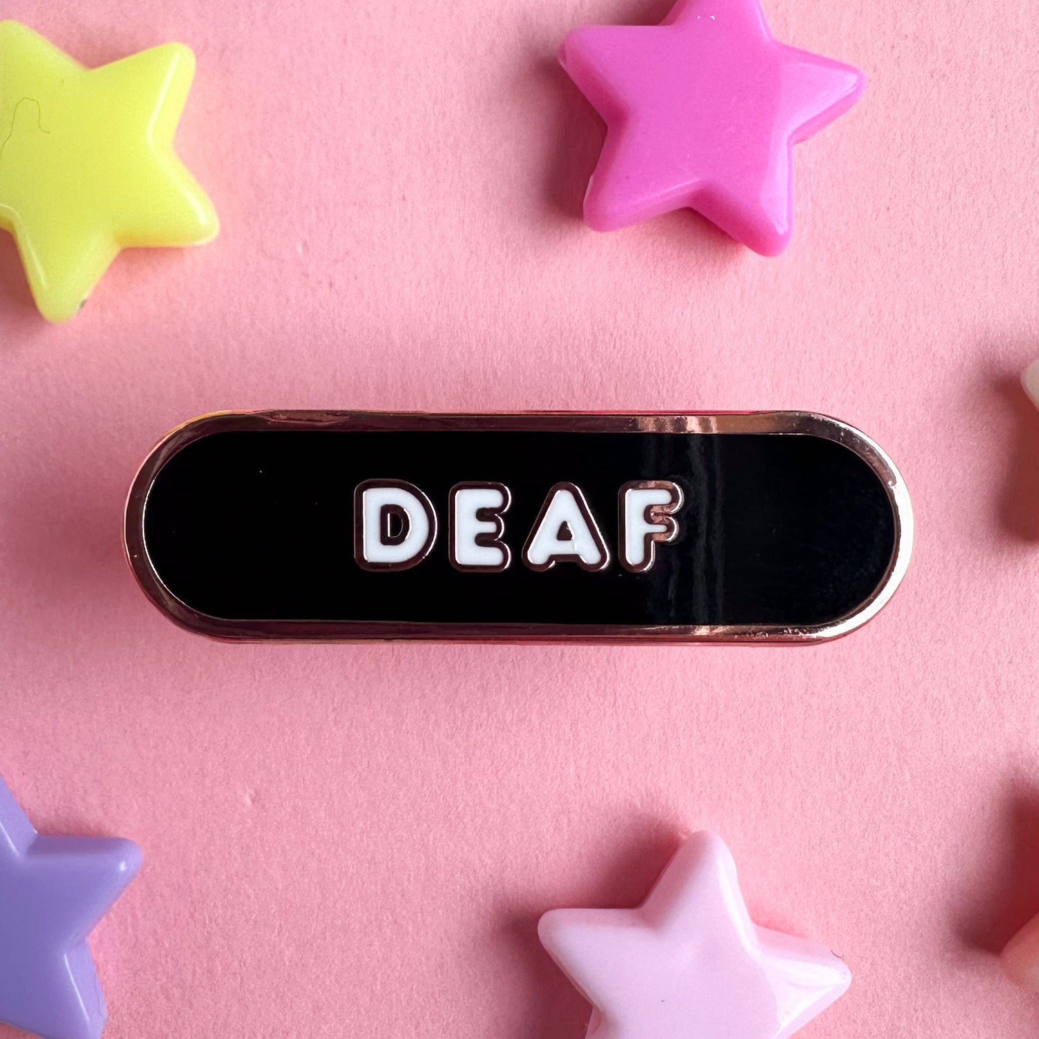 An oval shaped enamel pin that is black with white bubble letters that read "Deaf". The pin is on a pink background with plastic star bead around it. 