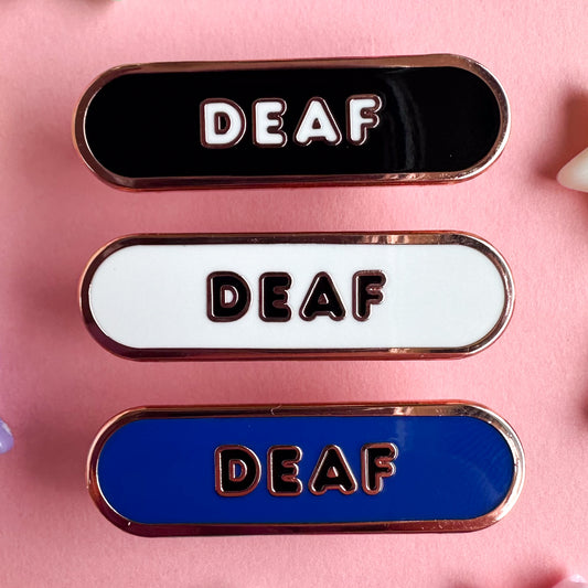 Three capsule shaped enamel pins that all have bubble letters that read "Deaf". The top pin is black the middle pin is white, the bottom pin is blue. The pins are on a pastel pink background.