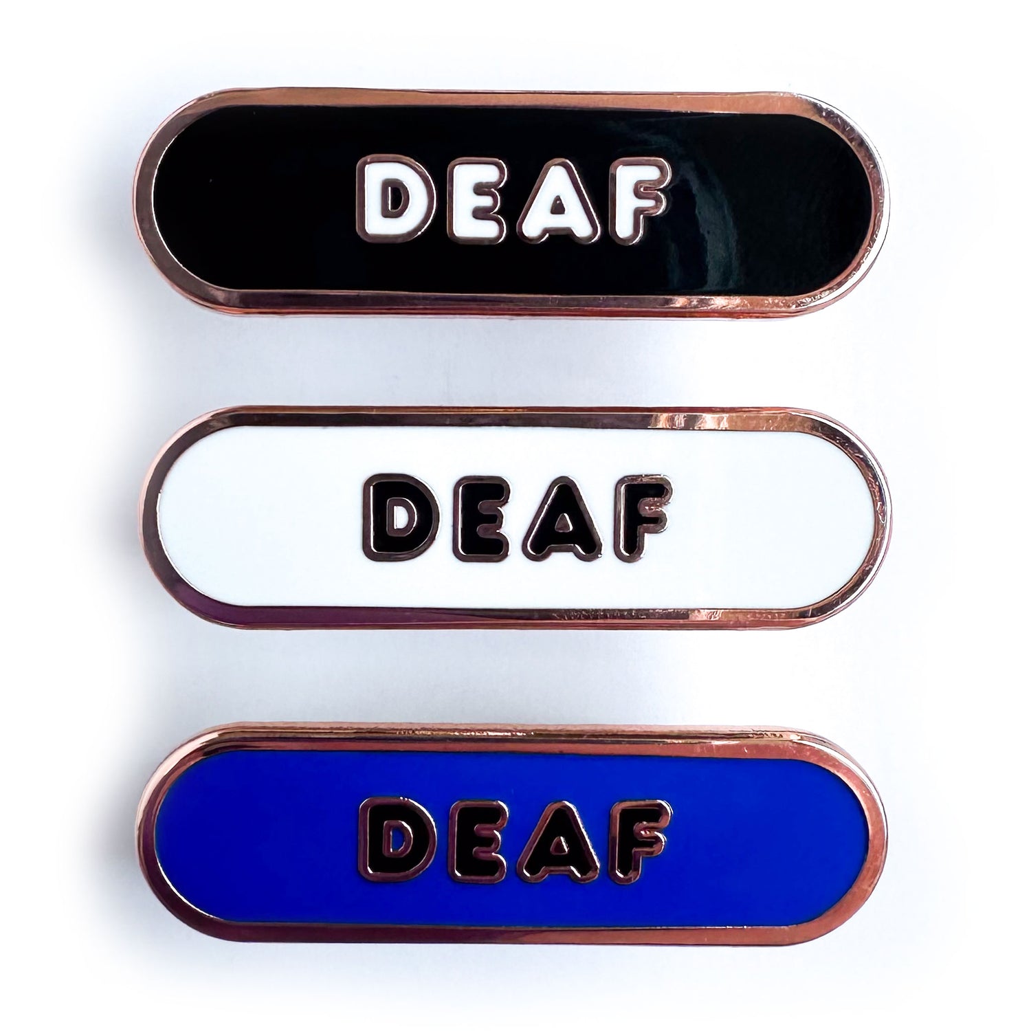 Three capsule shaped enamel pins that all have bubble letters that read "Deaf". The top pin is black the middle pin is white, the bottom pin is blue.