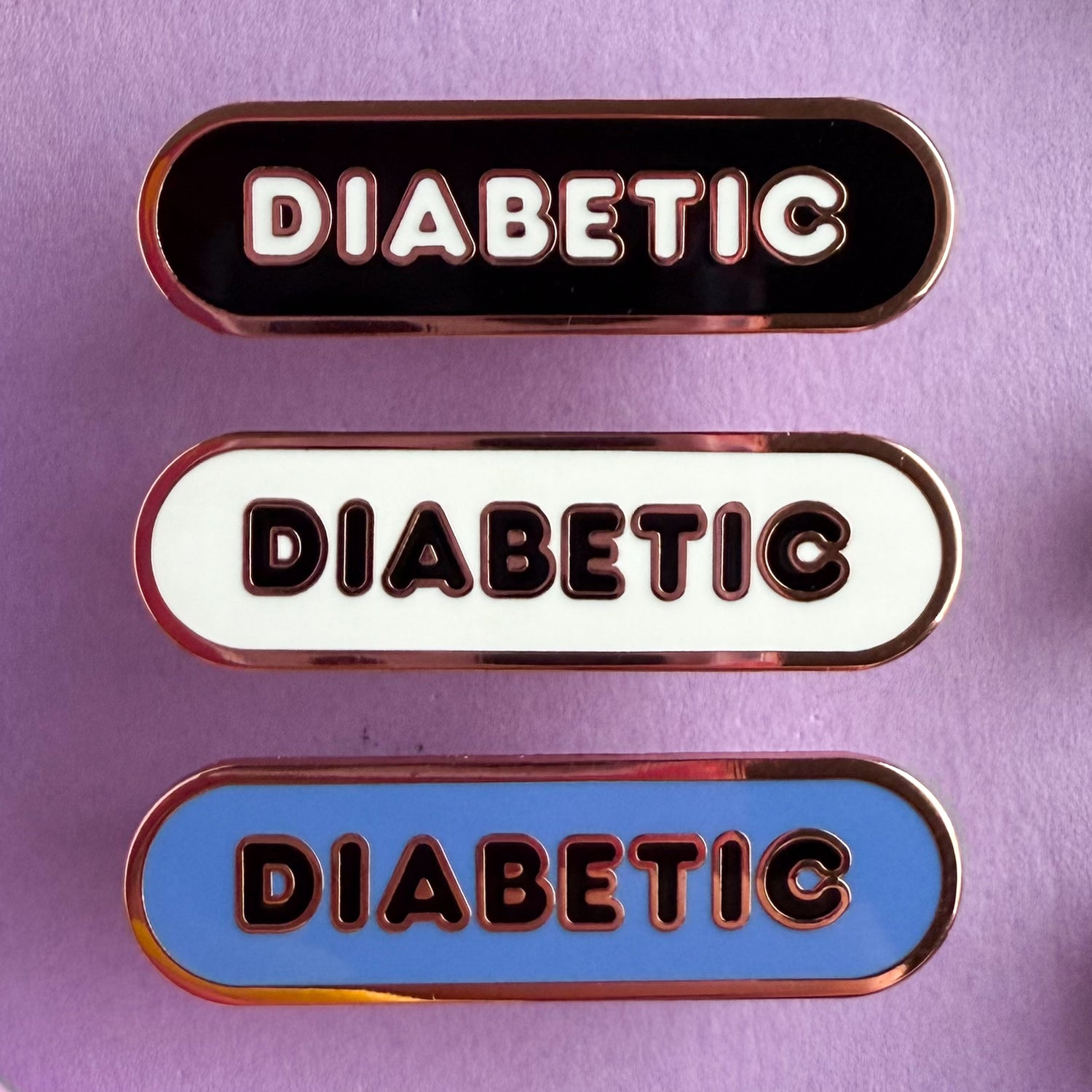 Three bandaid shaped pins with the text DIABETIC in bubble letters. Top one black with white text, the next is white with black text, and the last is cornflower blue with black text. The pins are on a purple background.