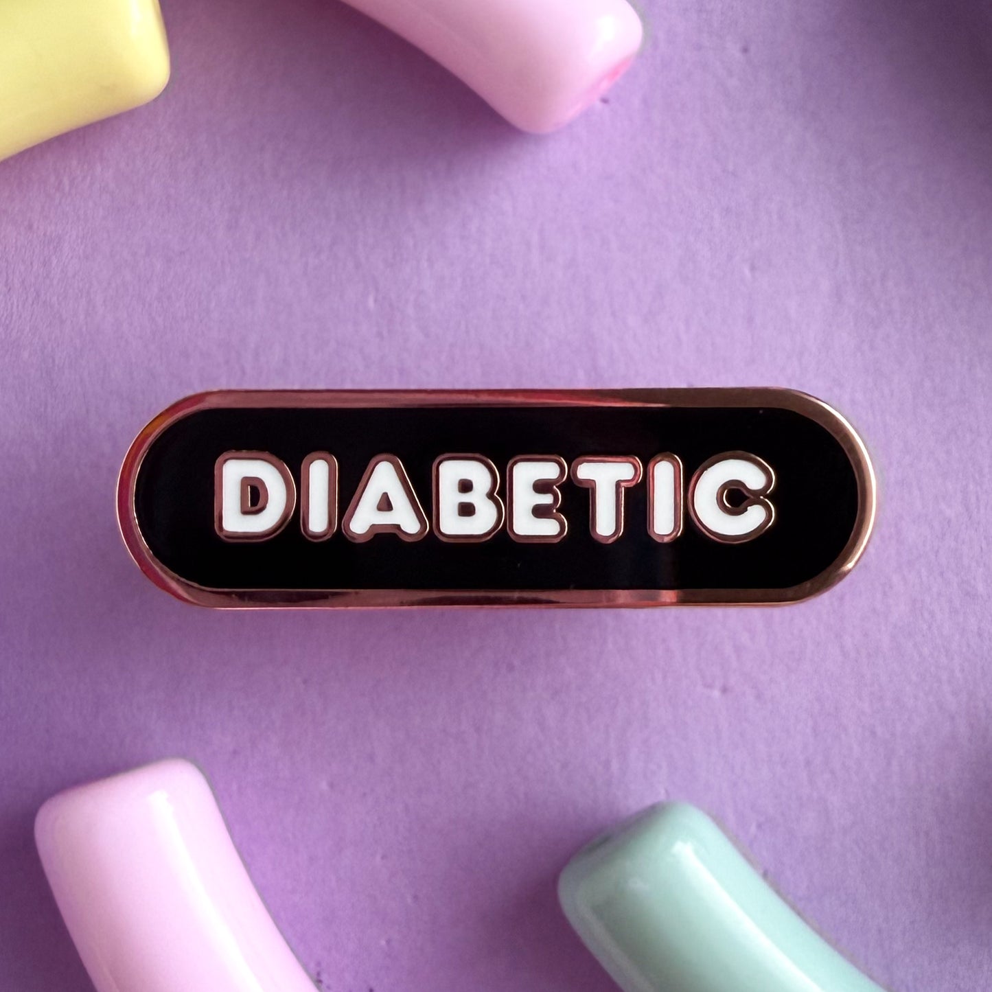 A bandaid shaped pin with the text DIABETIC in bubble letters. The background is black and the text is white. The pin is on a purple background.