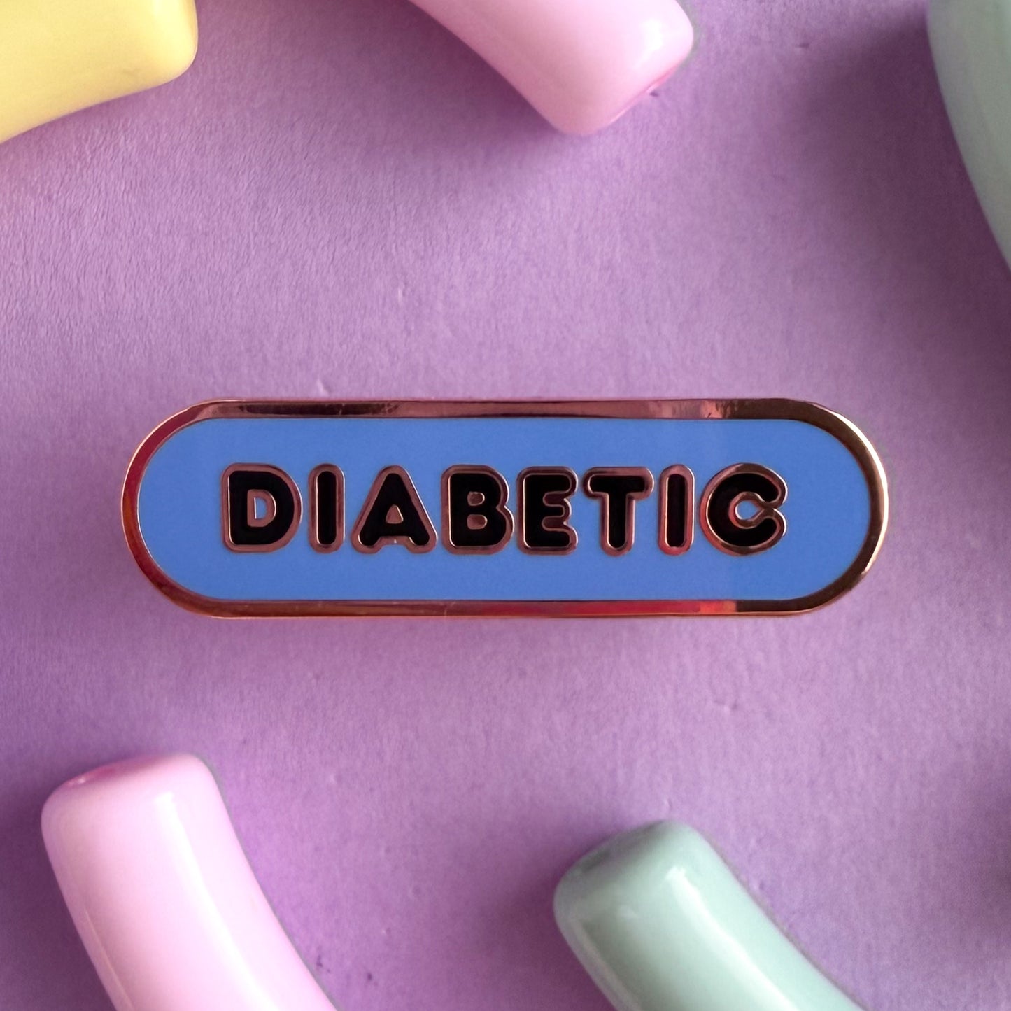 A bandaid shaped pin with the text DIABETIC in bubble letters. The background is cornflower blue and the text is black. The pin is on a purple background.
