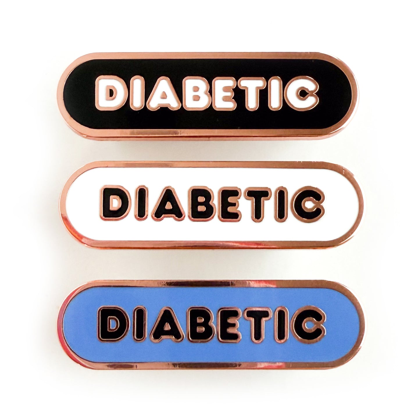 Three bandaid shaped pins with the text DIABETIC in bubble letters. Top one black with white text, the next is white with black text, and the last is cornflower blue with black text.