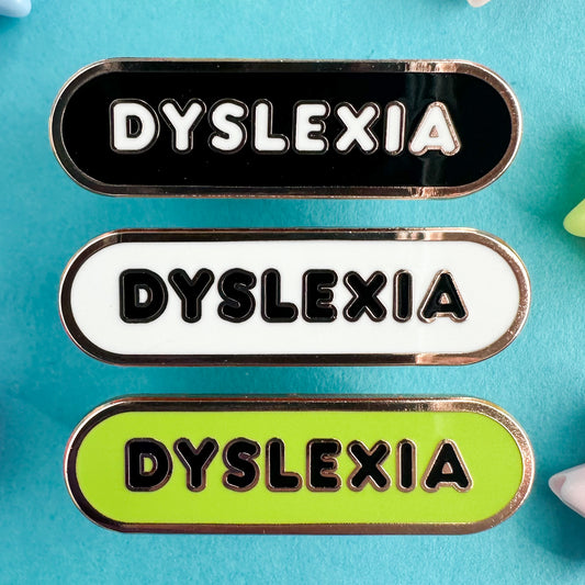 Three enamel pins in a stack that all read "Dyslexia" in bubble letters. The top pin is black, the middle is white and the bottom is lime green.  The pins are on a blue paper background.