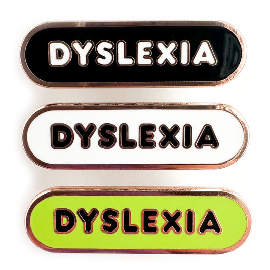 Three enamel pins in a stack that all read "Dyslexia" in bubble letters. The top pin is black, the middle is white and the bottom is lime green. 