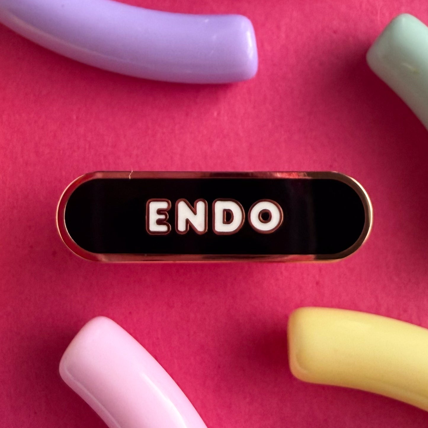A bandaid shaped pin with the text ENDO in bubble letters. The background is black and the text is white. The pin is on a pink background.