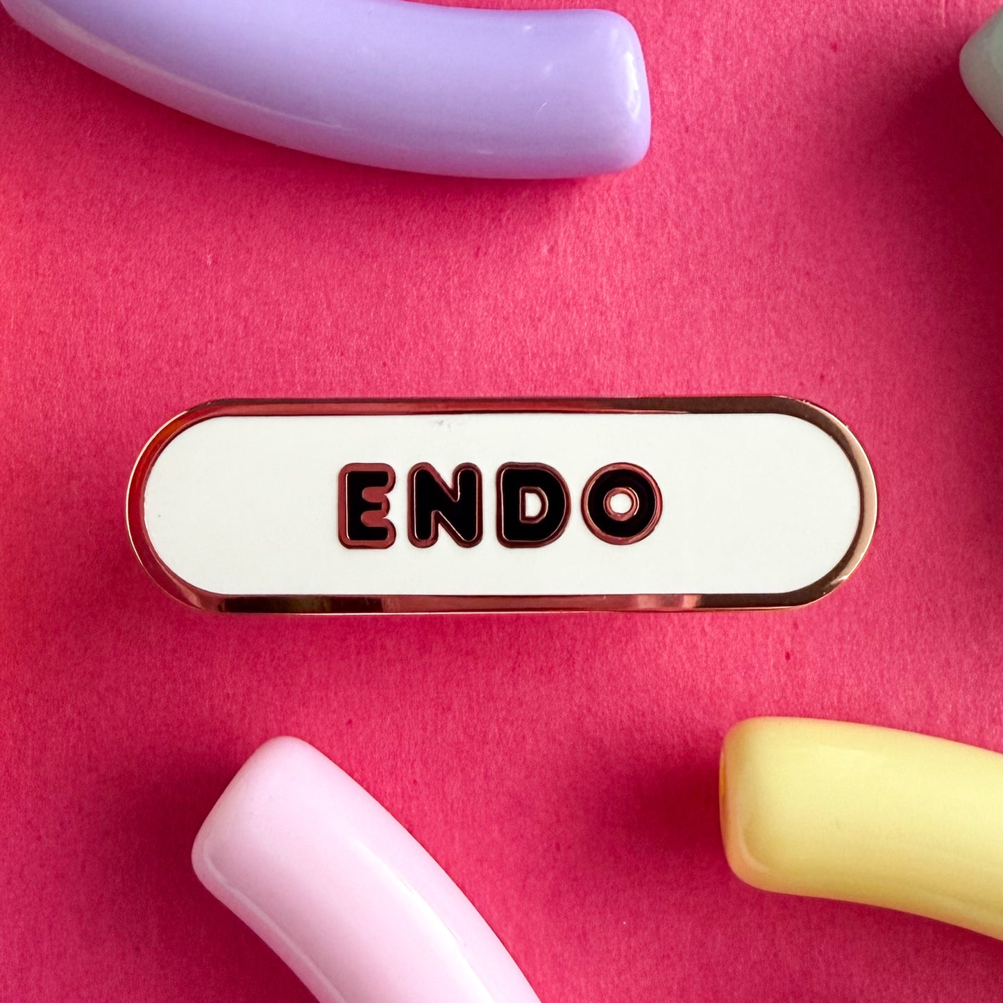 A bandaid shaped pin with the text ENDO in bubble letters. The background is white and the text is black. The pin is on a pink background.