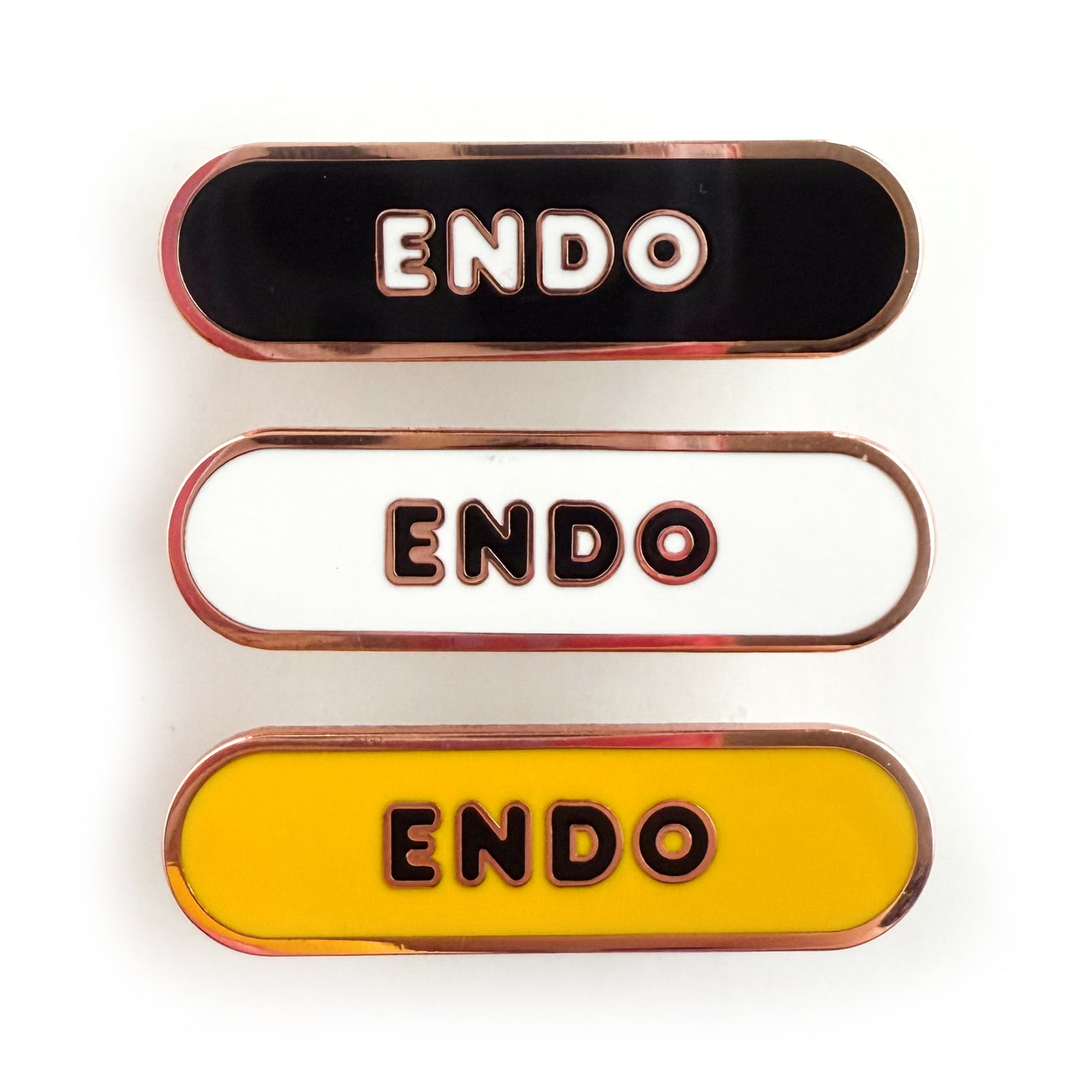 Three bandaid shaped pins with the text ENDO in bubble letters. Top one black with white text, the next is white with black text, and the last is yellow with black text.