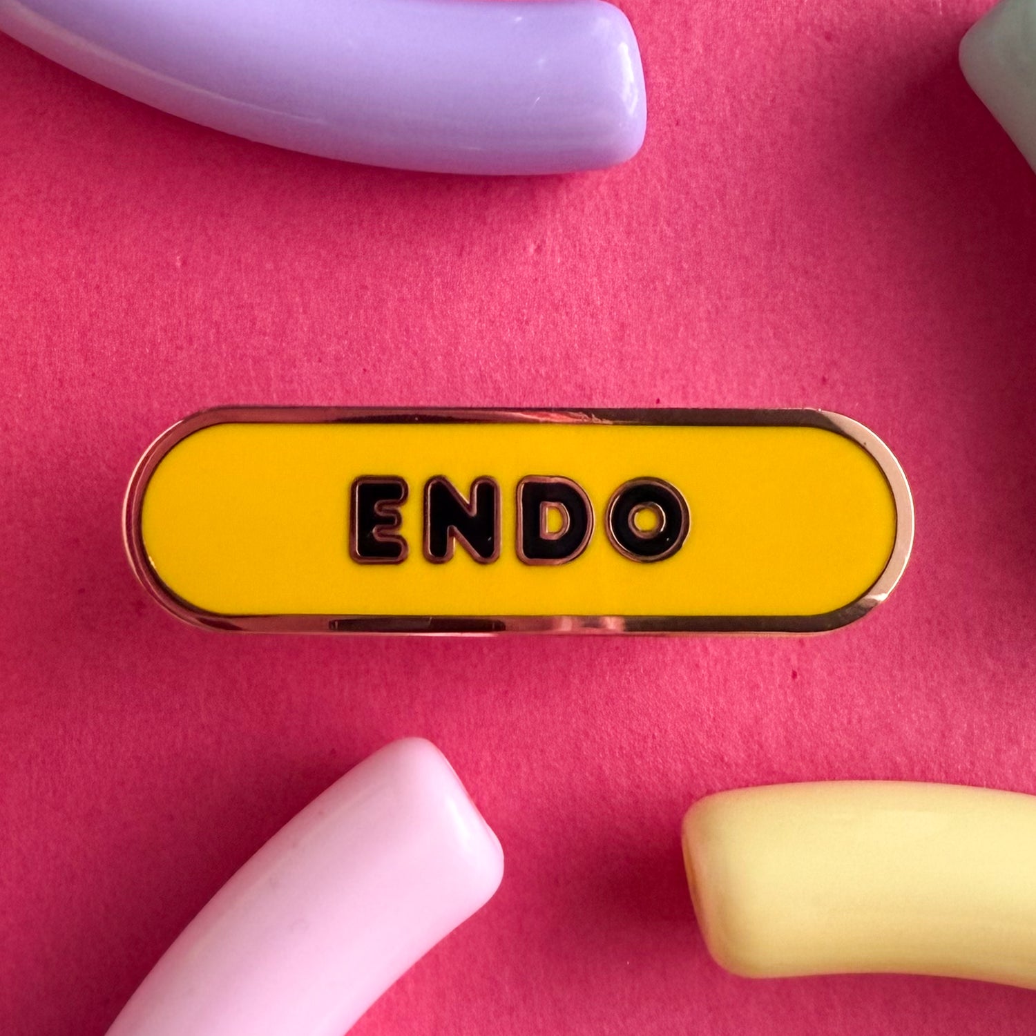 A bandaid shaped pin with the text ENDO in bubble letters. The background is yellow and the text is black. The pin is on a pink background.