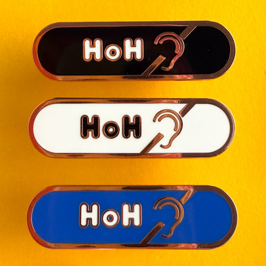Three bandaid shaped pins with the text HoH in bubble letters and the hard of hearing symbol. Top one black with white text, the next is white with black text, and the last is blue with white text. The pins are on a yellow background.