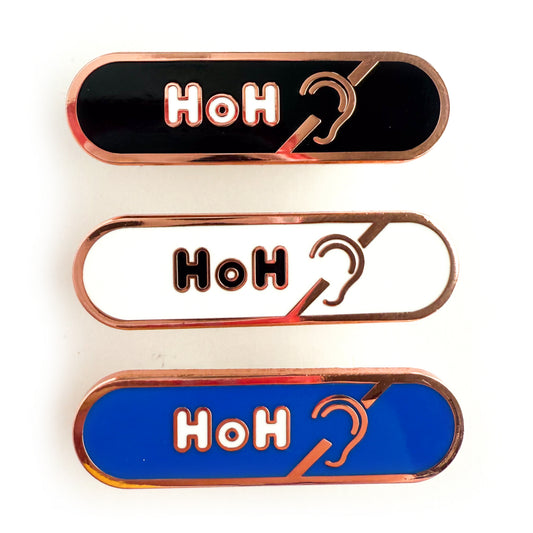 Three bandaid shaped pins with the text HoH in bubble letters and the hard of hearing symbol. Top one black with white text, the next is white with black text, and the last is blue with white text.