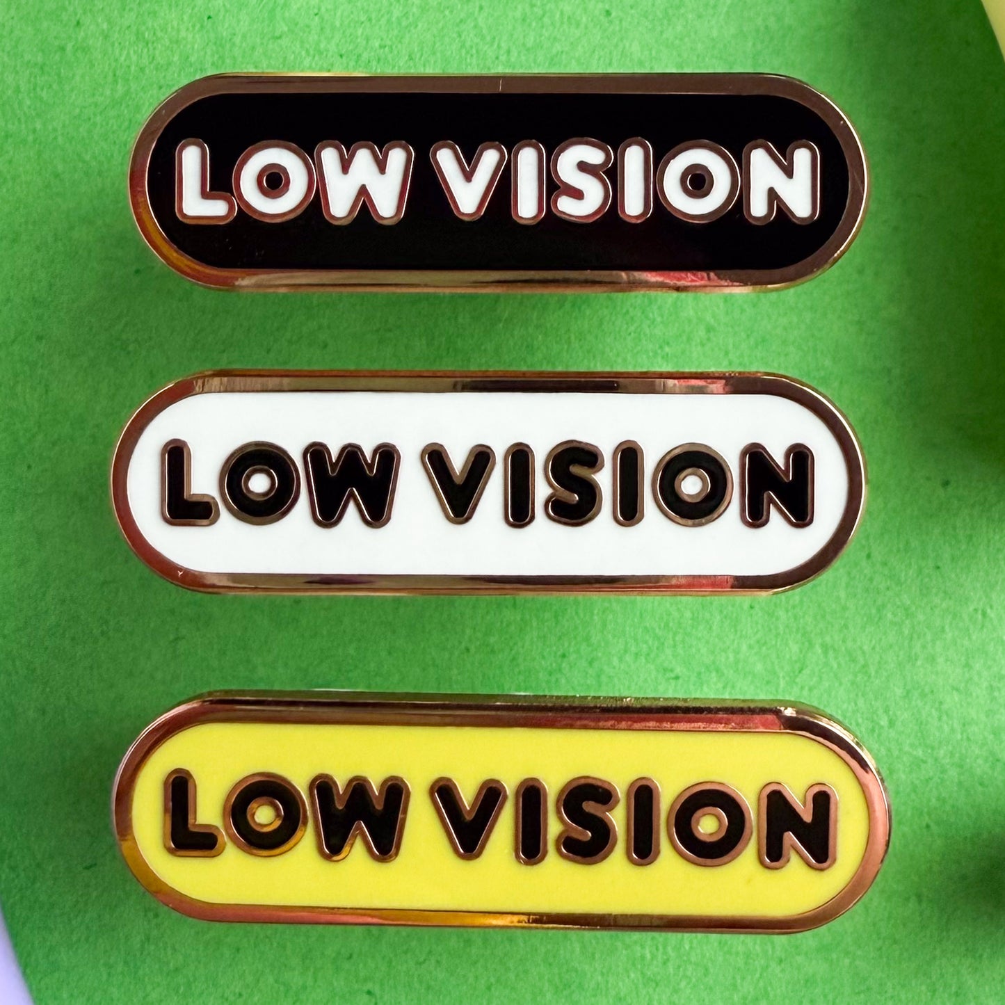 Three bandaid shaped pins with the text LOW VISION in bubble letters. Top one black with white text, the next is white with black text, and the last is yellow with black text. The pins are on a green background.