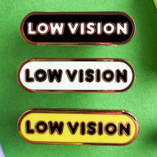Three bandaid shaped pins with the text LOW VISION in bubble letters. Top one black with white text, the next is white with black text, and the last is yellow with black text. The pins are on a green background.