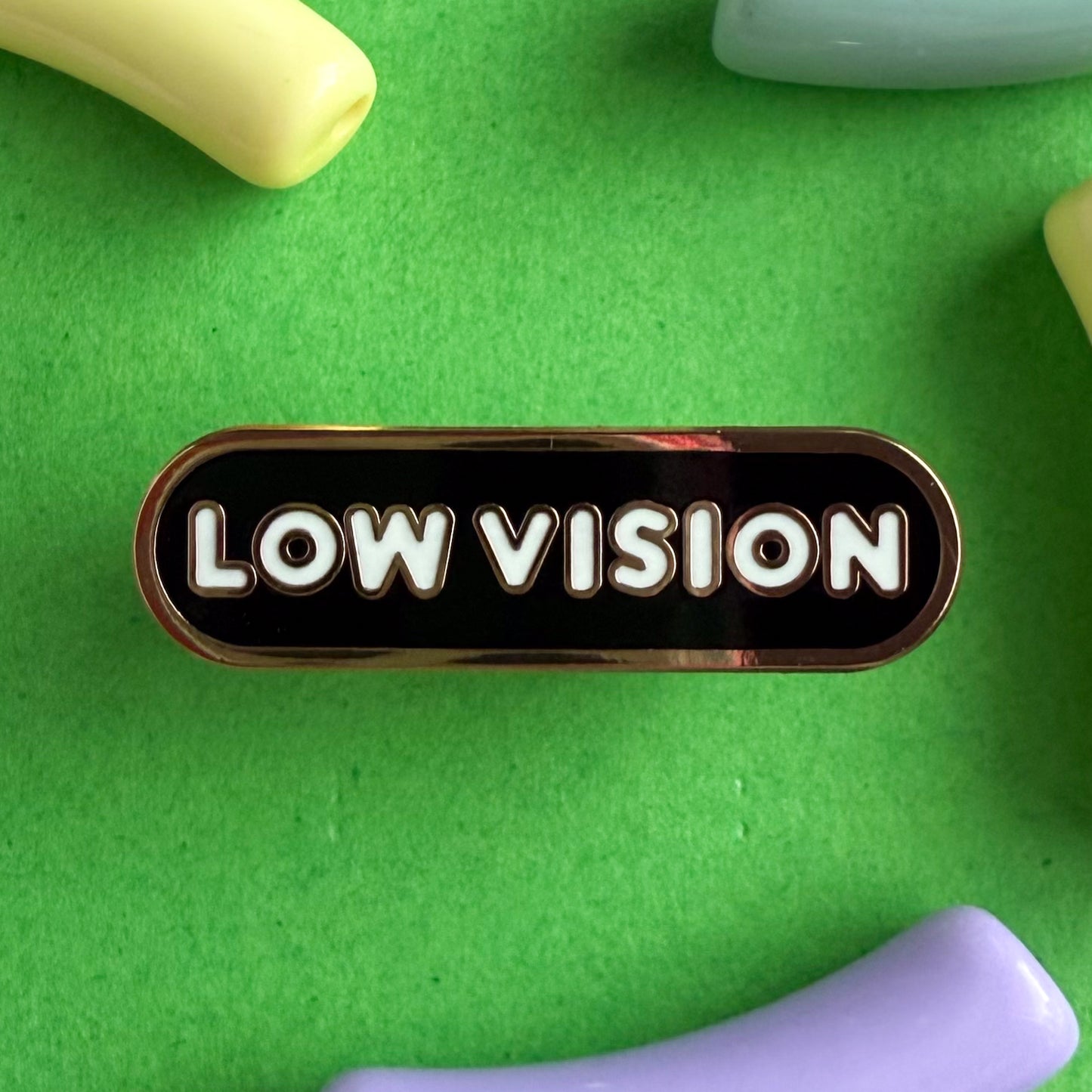A bandaid shaped pin with the text LOW VISION  in bubble letters. The background is black and the text is white. The pin is on a green background.