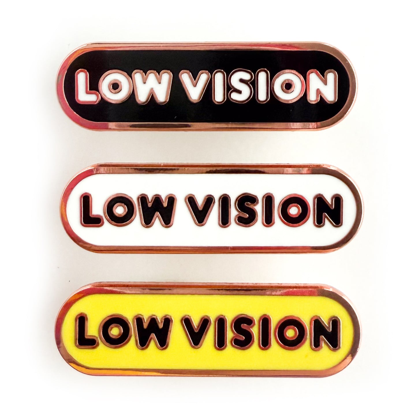 Three bandaid shaped pins with the text LOW VISION in bubble letters. Top one black with white text, the next is white with black text, and the last is yellow with black text.