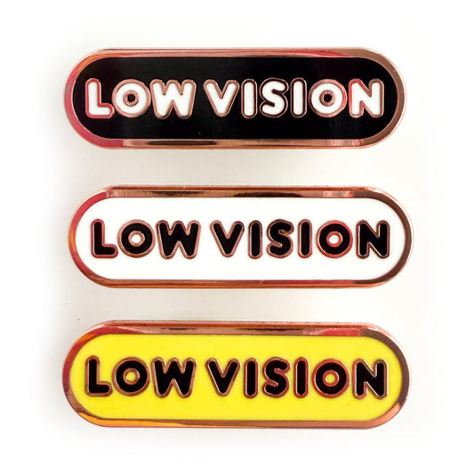 Three bandaid shaped pins with the text LOW VISION in bubble letters. Top one black with white text, the next is white with black text, and the last is yellow with black text.