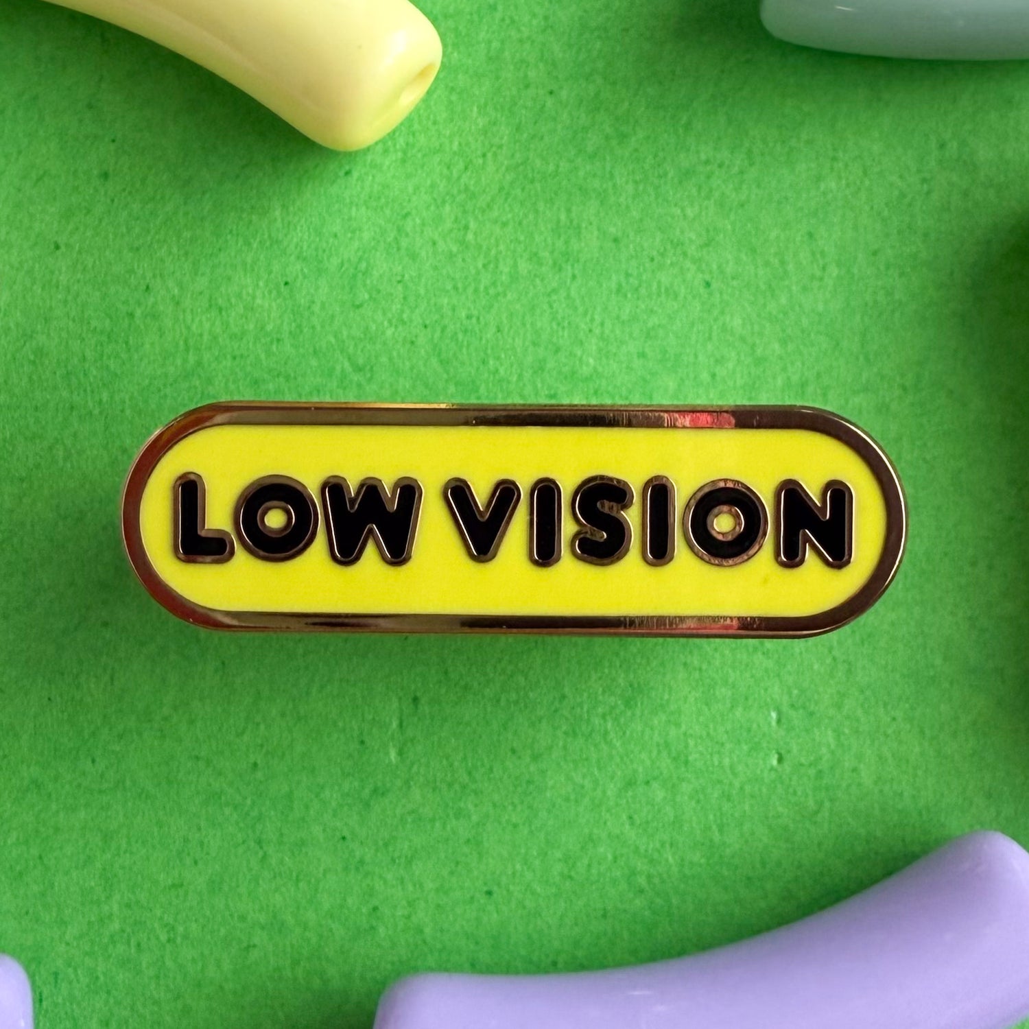 A bandaid shaped pin with the text LOW VISION  in bubble letters. The background is yellow and the text is black. The pin is on a green background.