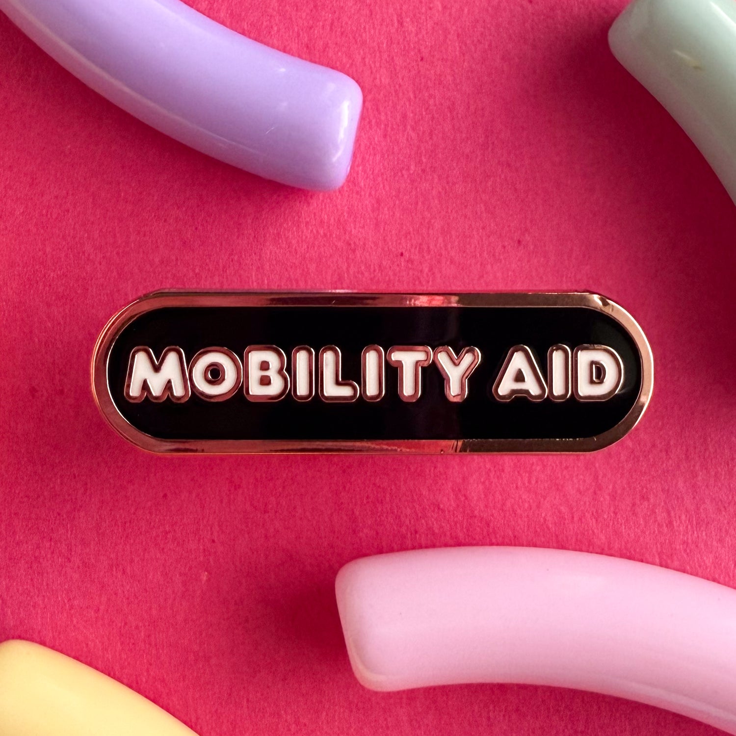 A bandaid shaped pin with the text MOBILITY AID in bubble letters. The background is black and the text is white. The pin is on a pink background.