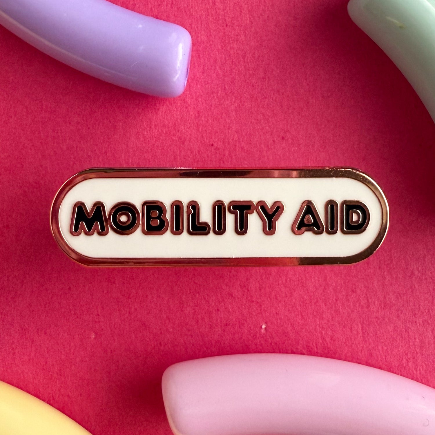 A bandaid shaped pin with the text MOBILITY AID  in bubble letters. The background is white and the text is black. The pin is on a pink background.