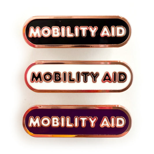 Three bandaid shaped pins with the text MOBILITY AID in bubble letters. Top one black with white text, the next is white with black text, and the last is eggplant purple with white text.