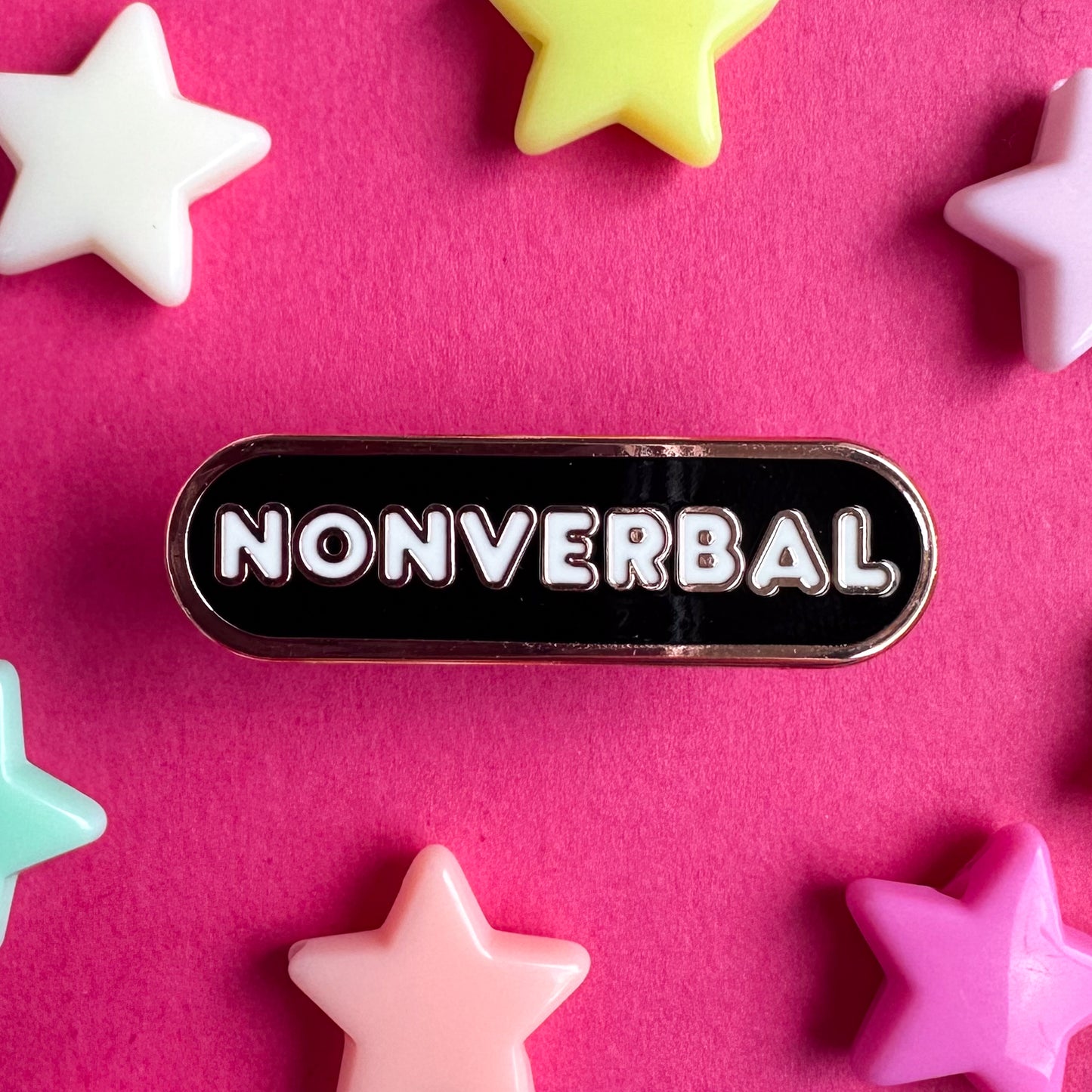 A capsule shaped enamel pin that has white bubble letters that read "Nonverbal" the majority of the pin is black with copper color metal. The pin is on a pink background with pastel star beads around it. 