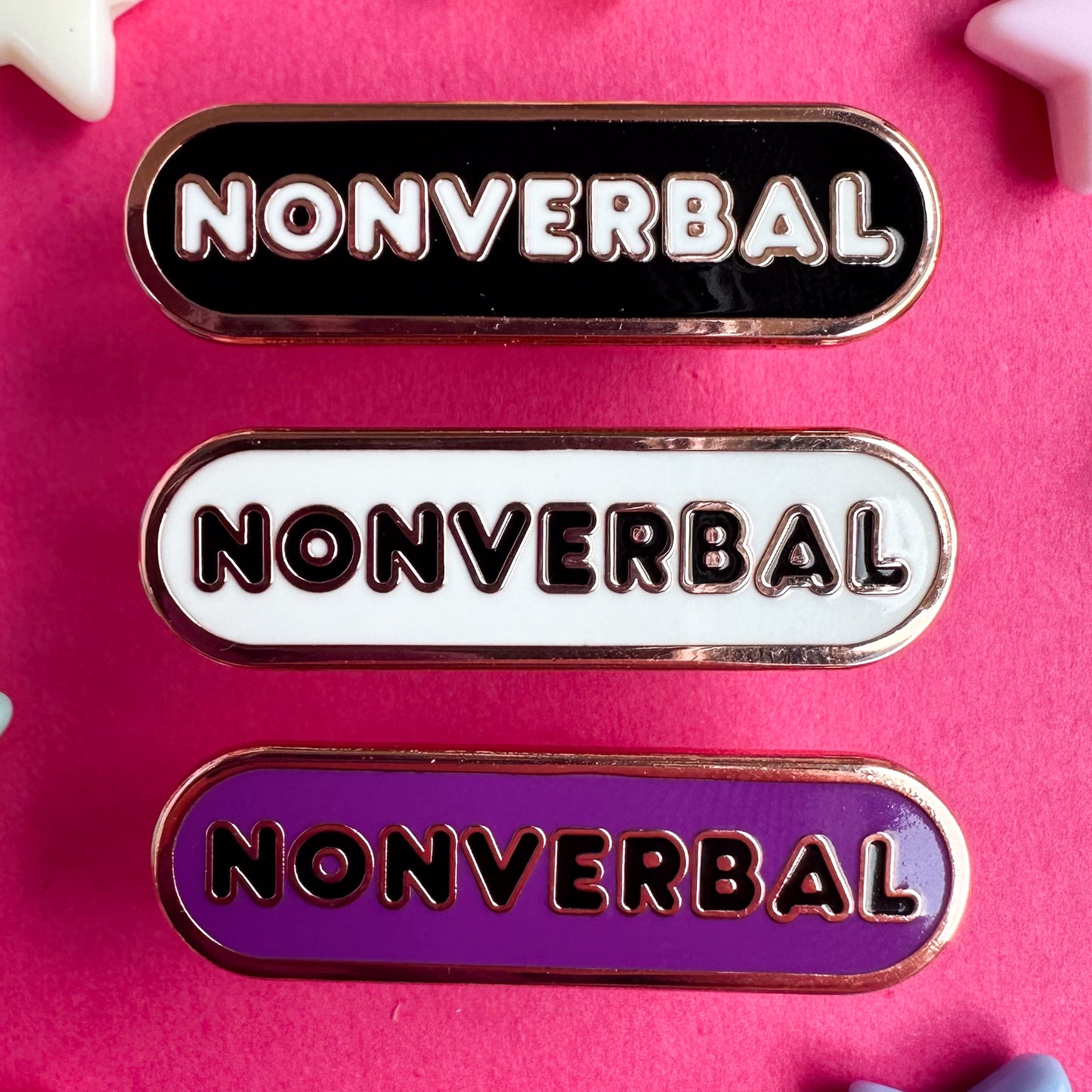 Three capsule shaped enamel pins that all have bubble letters that read "Nonverbal", the top pin is black, the middle is white, and the bottom is purple.  The pins are on a hot pink paper background.