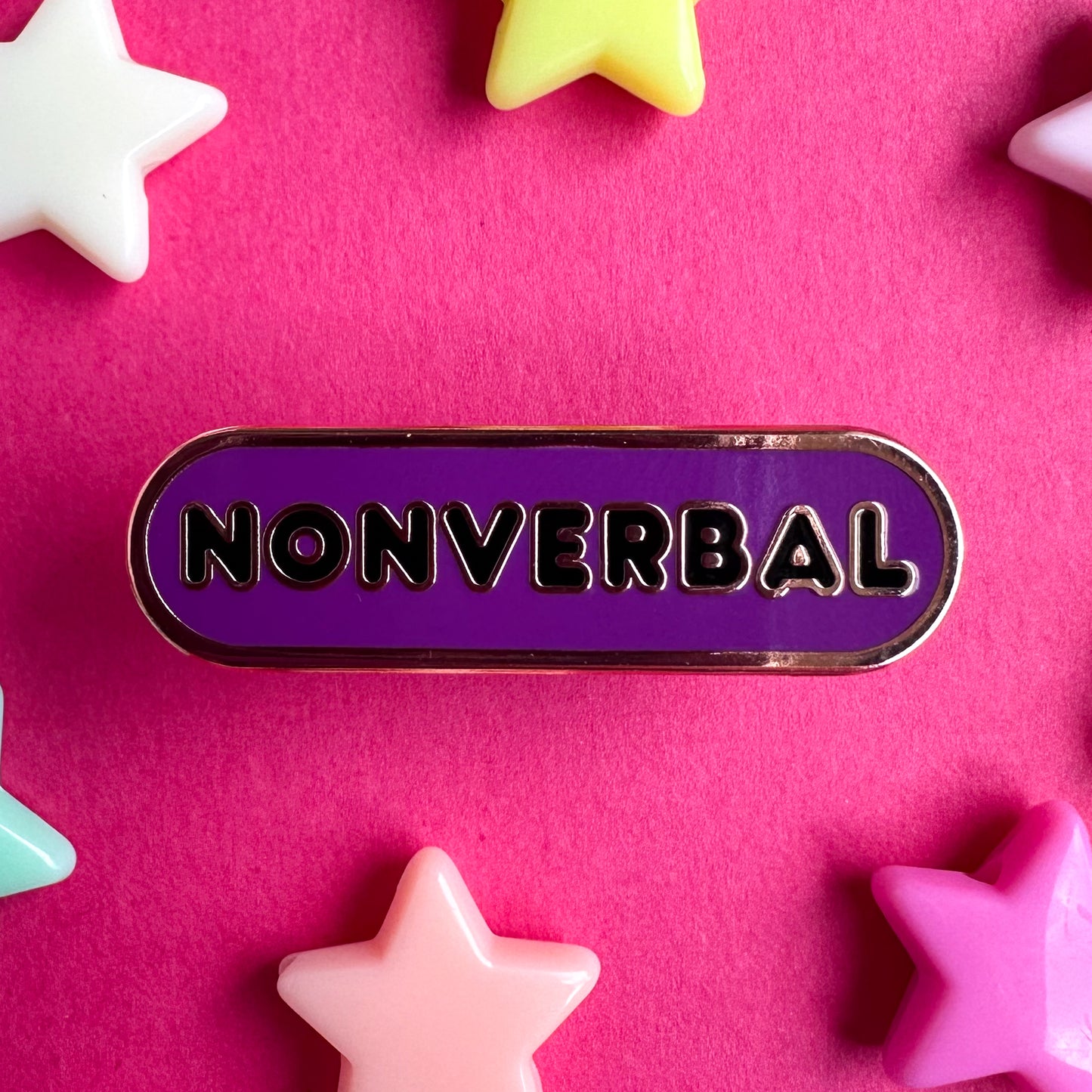 A capsule shaped pin that is purple with black bubble letters that spell out "Nonverbal". The pin is on a hot pink paper background with plastic star beads around it. 