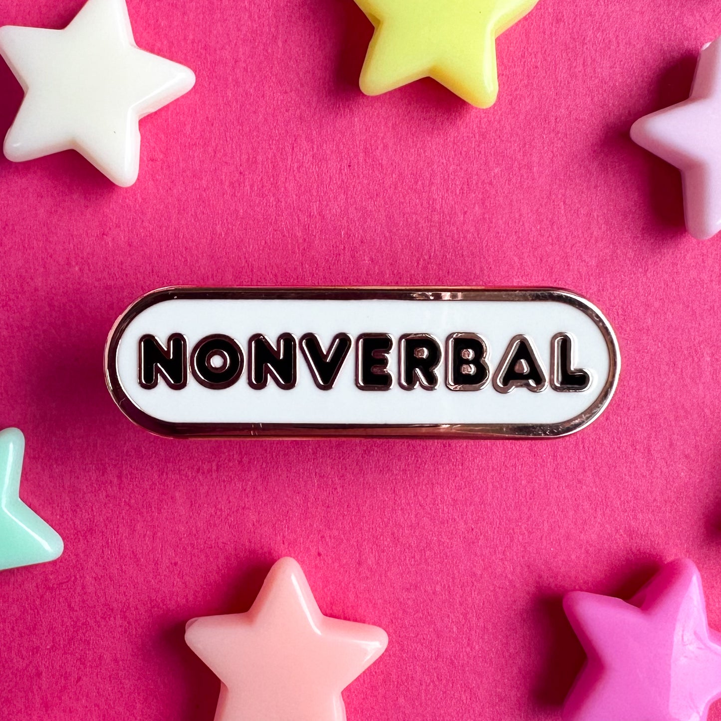 A white oval shaped enamel pin that reads "Nonverbal". The pin is sitting on a hot pink background with plastic star beads around it. 