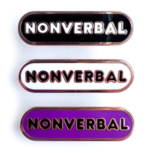 Three capsule shaped enamel pins that all have bubble letters that read "Nonverbal", the top pin is black, the middle is white, and the bottom is purple.