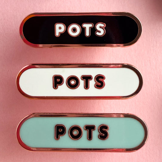 Three bandaid shaped pin with the text POTS in bubble letters. Top one black with white text, the next is white with black text, and the last is light blue with black text. The pins are on a pink background.