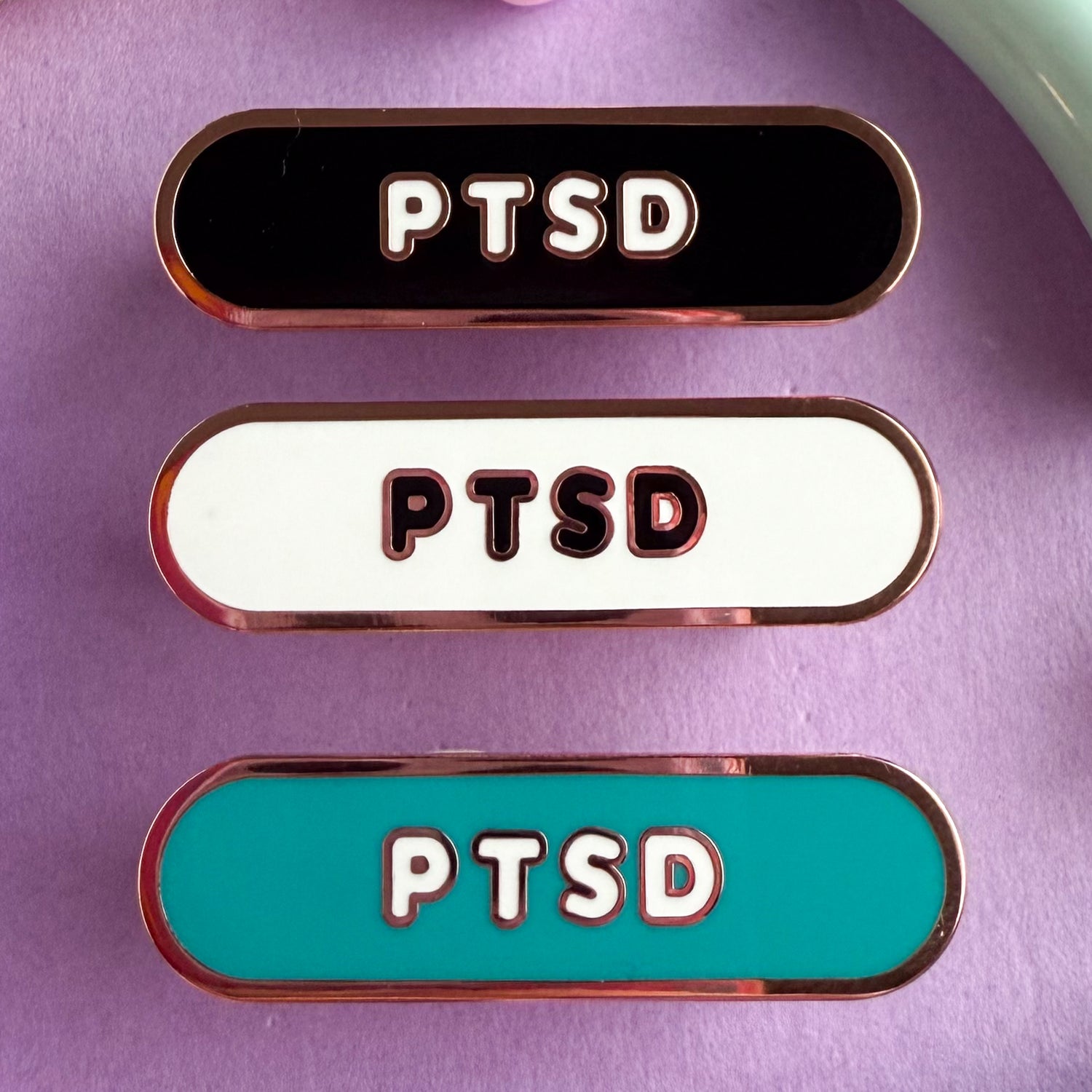 Three bandaid shaped pin with the text PTSD in bubble letters. Top one black with white text, the next is white with black text, and the last is teal with white text. These pins are on a purple background.