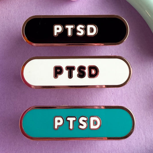 Three bandaid shaped pin with the text PTSD in bubble letters. Top one black with white text, the next is white with black text, and the last is teal with white text. These pins are on a purple background.