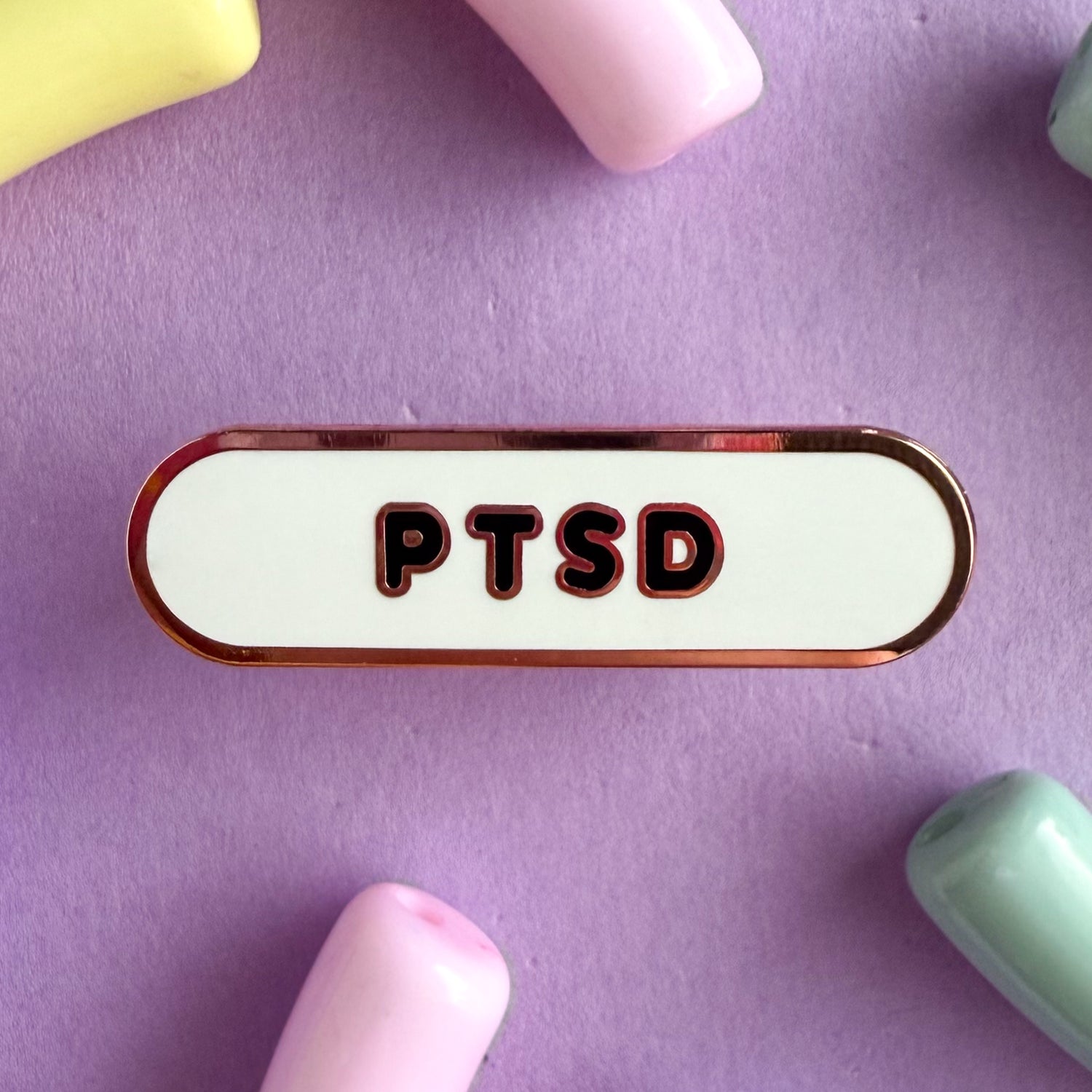 A bandaid shaped pin with the text PTSD in bubble letters. The background is white and the text is black. The pin is on a purple background.