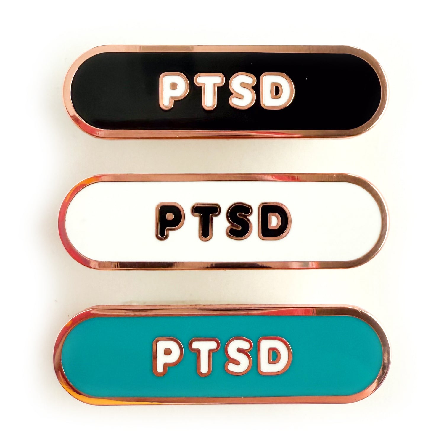 Three bandaid shaped pins with the text PTSD in bubble letters. Top one black with white text, the next is white with black text, and the last is teal with white text.