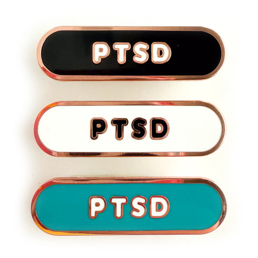 Three bandaid shaped pins with the text PTSD in bubble letters. Top one black with white text, the next is white with black text, and the last is teal with white text.
