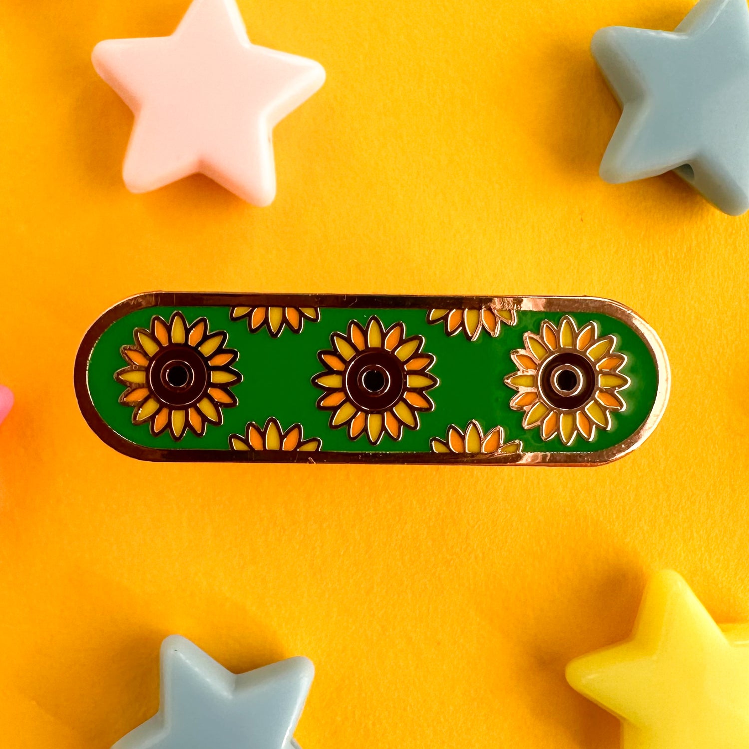 A capsule shaped pin with illustrations of sunflowers on a green background representing invisible disabilities. The pin is on a yellow backdrop with pastel star beads strewn around it. 