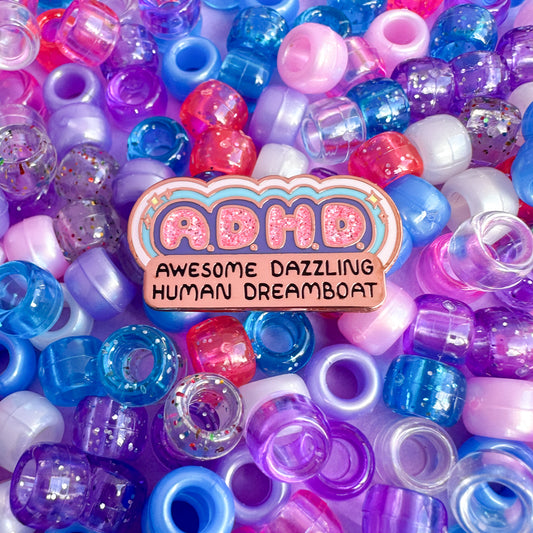 An enamel pin that reads A.D.H.D. Awesome Dazzling Human Dreamboat sitting on top of a pile of glitter pony beads in purple, blue, and pink, which are the same colors of the pin.