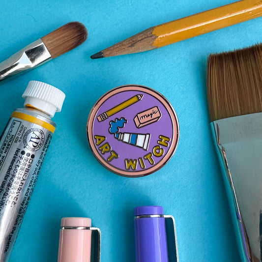 A merit b badge style pin that reads "Art Witch" with art supplies on the pin surrounded by real art supplies like a pencil, paint tube, and paint brushes on a blue paper background.