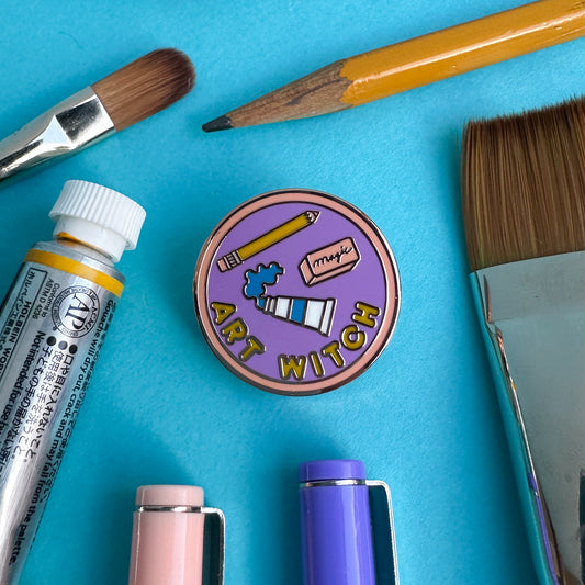 A merit b badge style pin that reads "Art Witch" with art supplies on the pin surrounded by real art supplies like a pencil, paint tube, and paint brushes on a blue paper background. 