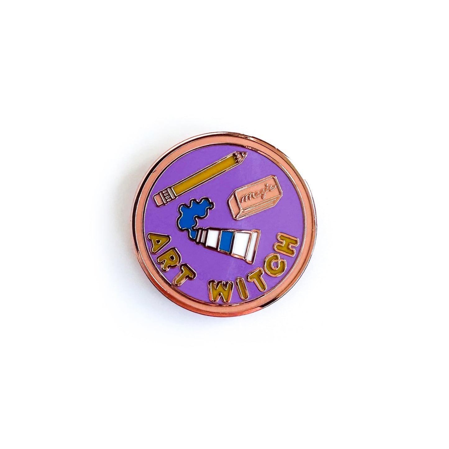 A circular enamel pin that has "Art Witch" on it in bubble letters with images of a paint tube, a pencil and a pink eraser on it. The background of the pin is purple with a pink border. 