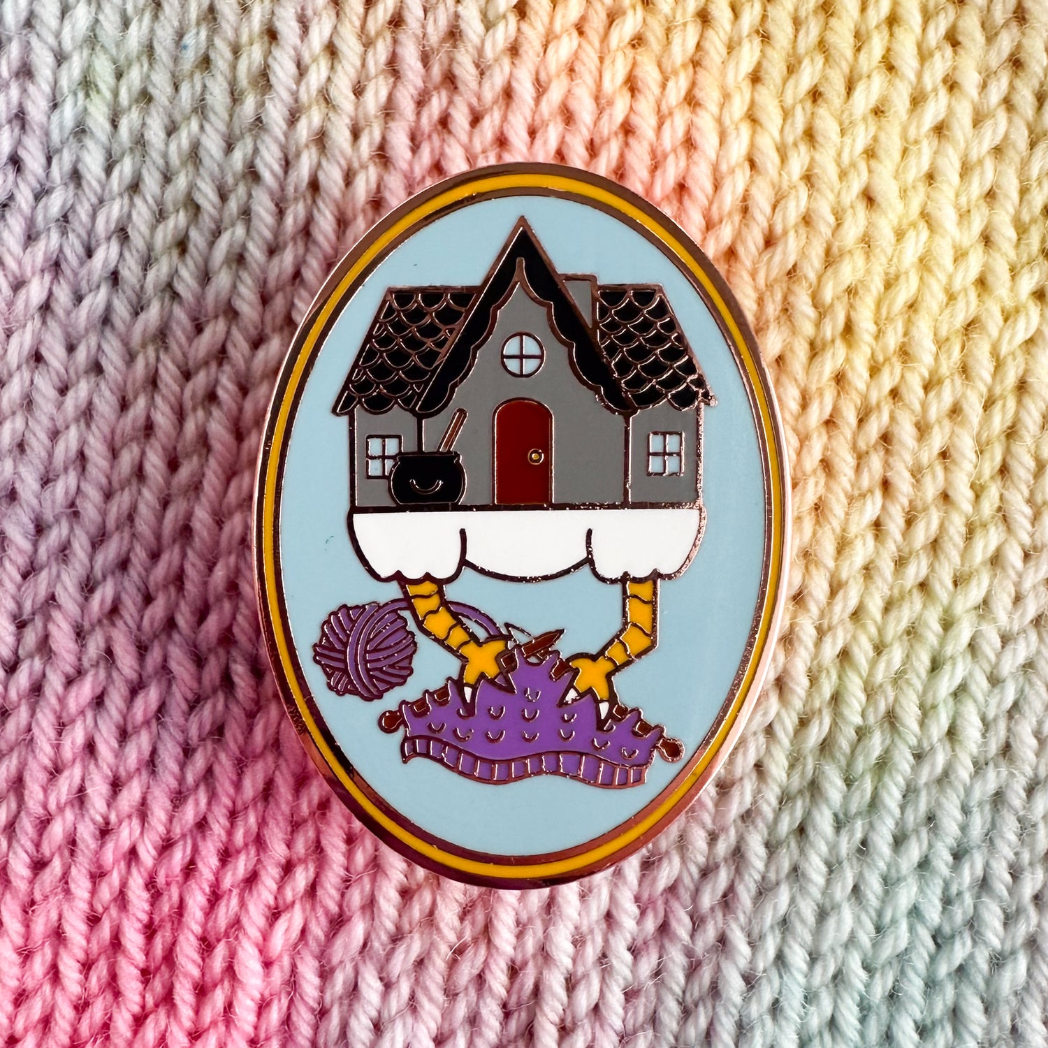 An oval shaped enamel pin with a cartoon illustration of Baba Yaga's house knitting on a purple knitting project with its chicken feet. The pin has a blue background with a yellow border and is sitting on a pastel stockinette stitch background. 