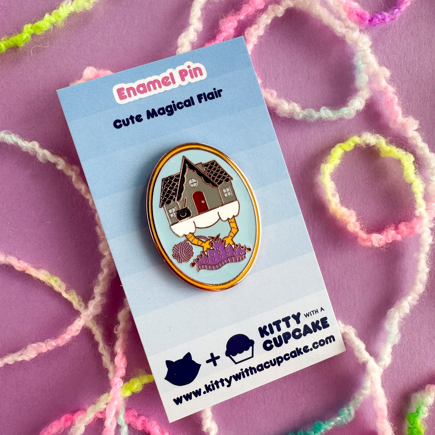 An enamel pin the shape of an oval with an illustration of Baba Yaga's house with its chicken feet knitting with purple yarn. The pin is packaged on a blue card that read "Enamel Pin - Cute Magical Flair" at the top. The card is sitting on a purple paper backdrop with pastel yarn strewn around it. 