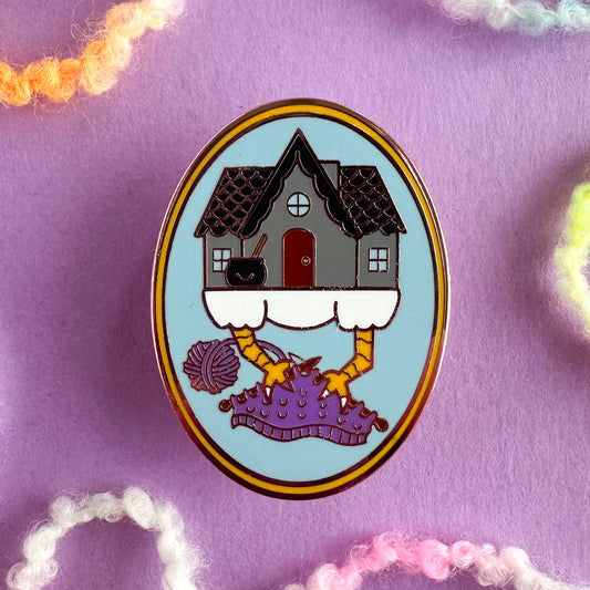 An oval shaped enamel pin with a cute illustration of Baba Yaga's house knitting on a project with its chicken feet. The oval is light blue with a yellow border. The house is grey with a black roof and the yarn of the knitting project is purple. The pin is on a purple paper background with bits of yarn strewn around it. 