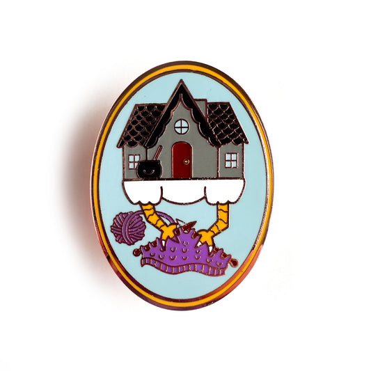 An oval shaped enamel pin with a cute illustration of Baba Yaga's house knitting with its chicken feet. The knitting project is purple and has a purple yarn ball attached.