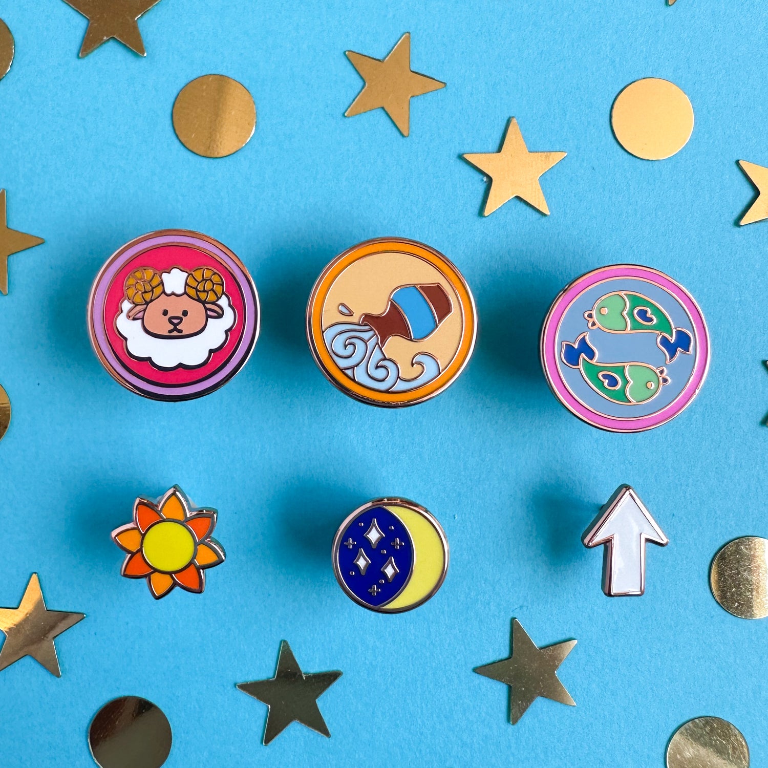 A set of six enamel pins, three are circles with cute illustrations representing zodiac signs. and the other three are shaped like a sun, moon, and rising arrow. The pins are on a blue background strewn with gold confetti shaped like stars and circles.