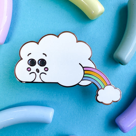 An enamel pin in the shape of a cloud with a face and a butt, there is a pastel rainbow arc coming out of the butt cheeks with another small cloud below it. The pin is on a blue background with pastel macaroni shaped beads around it.